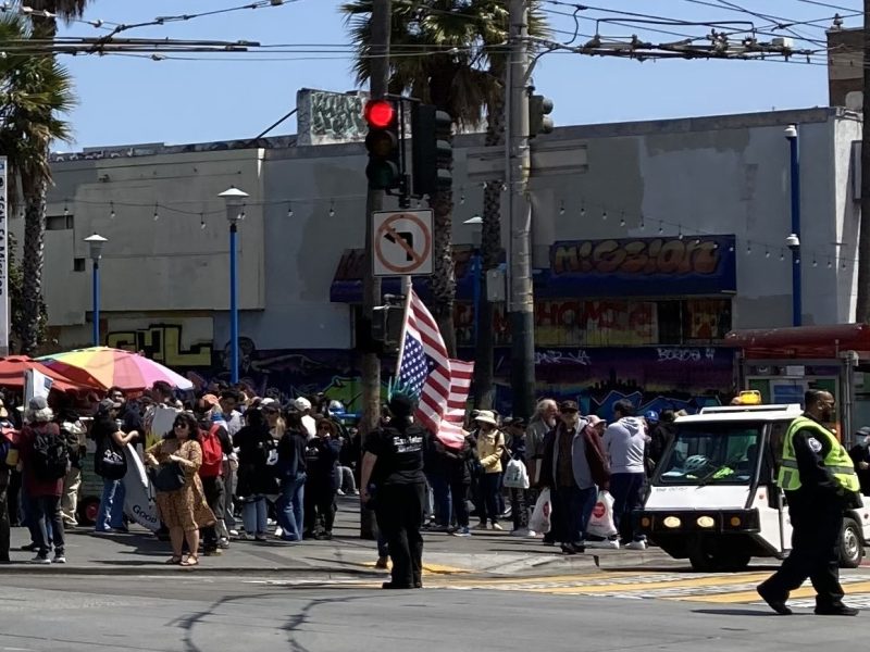 A crowd gathers at a street corner near a police officer, a small vehicle, and a person holding an American flag under sunny skies. Urban buildings and palm trees are in the background.