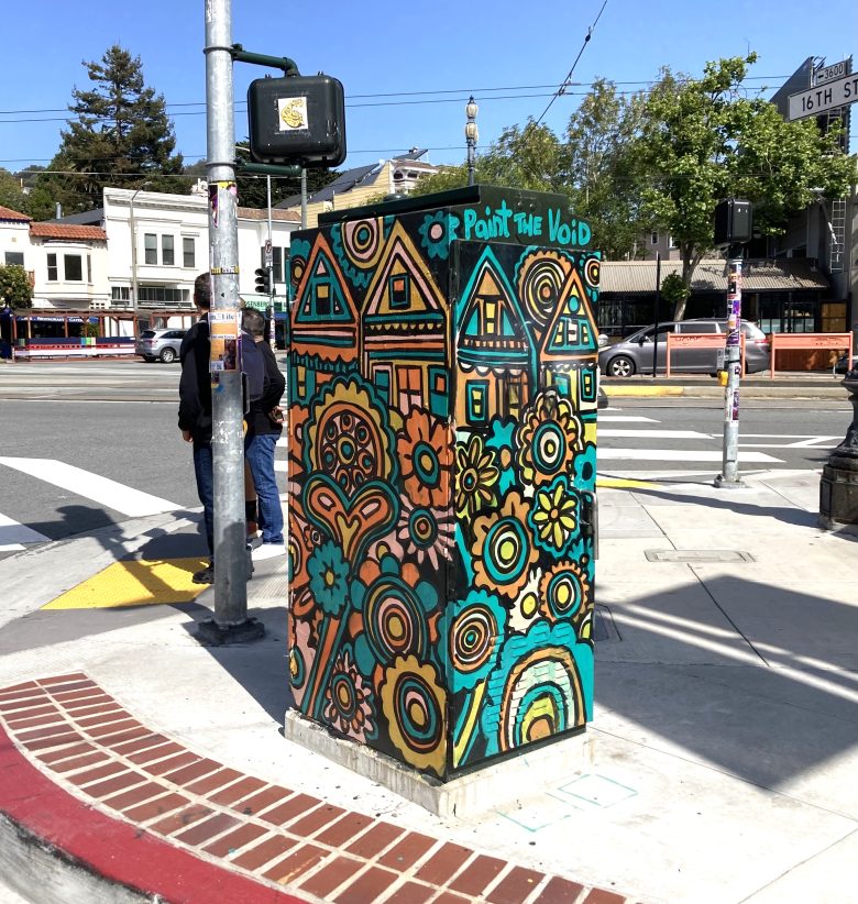 A utility box at a street corner painted with colorful flowers and houses, labeled “Paint the Void,” with buildings and pedestrians in the background.