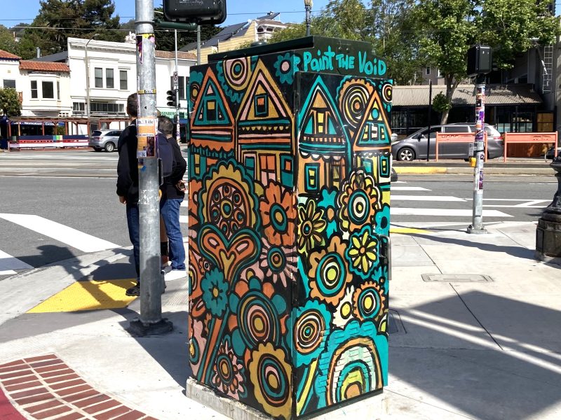 A utility box at a street corner painted with colorful flowers and houses, labeled “Paint the Void,” with buildings and pedestrians in the background.