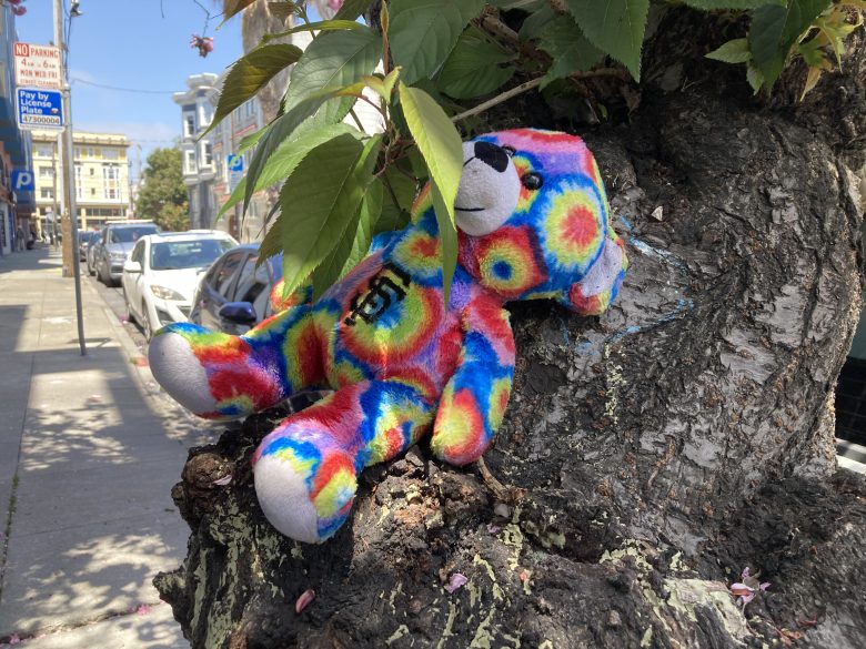 A colorful tie-dye teddy bear rests against the trunk of a tree on a city sidewalk, partially covered by leaves.