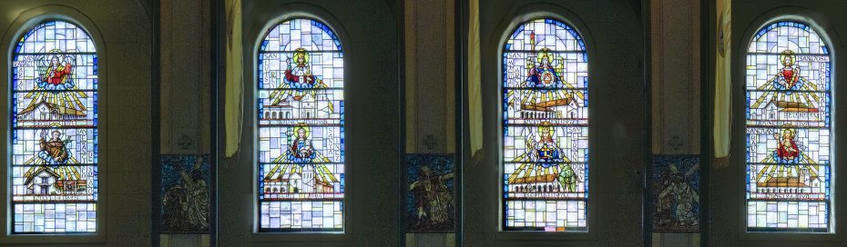 Four stained glass windows depict biblical figures and scenes, each with labeled names and intricate, colorful designs within arched frames inside a church.