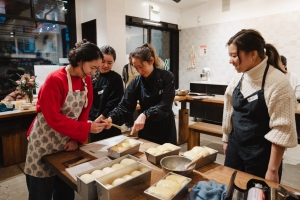 Four people wearing aprons prepare bread dough together at a wooden table in a kitchen setting.