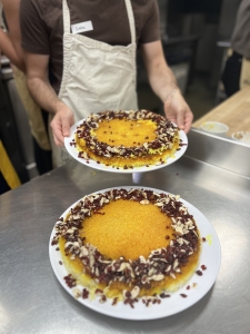 Person in an apron holding a plate with a round cake; another similar cake garnished with nuts and dried fruit sits on a counter in the foreground.