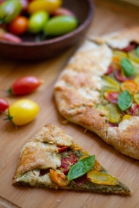 A slice of vegetable galette with basil leaves sits on a wooden surface next to the rest of the galette and a bowl of colorful tomatoes.