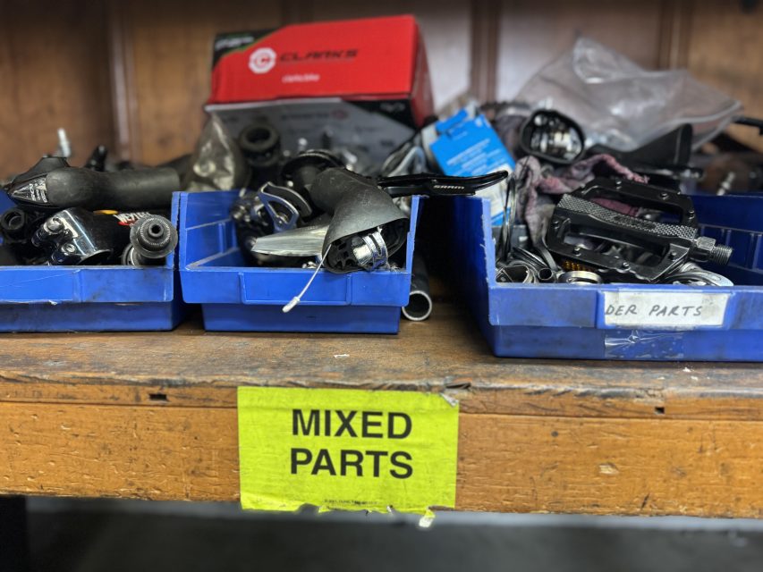 Three blue bins labeled "MIXED PARTS" on a wooden shelf, each containing assorted bicycle parts and components in a workshop setting.