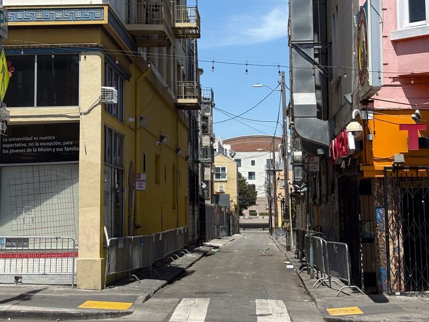 A narrow urban alley lined with yellow and pink buildings, barricades, string lights overhead, and debris scattered on the ground under a sunny sky.