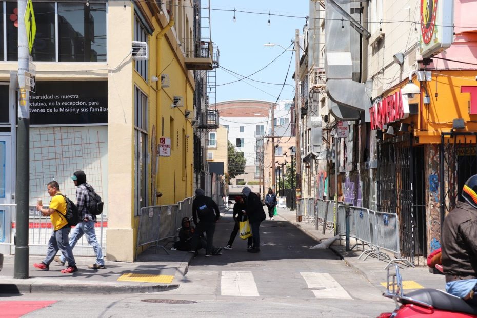 A narrow urban alleyway with several people walking or standing, bordered by yellow and tan buildings and temporary metal barriers on both sides.