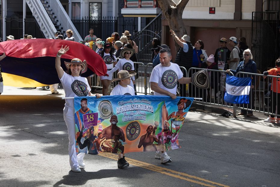 A group of people march in a parade holding a colorful banner and wearing matching white shirts and straw hats, with onlookers and a flag in the background.