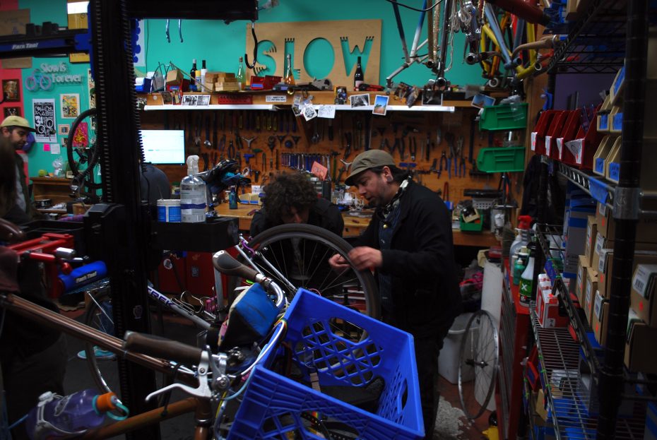 People repair bicycles in a cluttered bike workshop, surrounded by tools, parts, and equipment, with a large "SLOW" sign on the back wall.