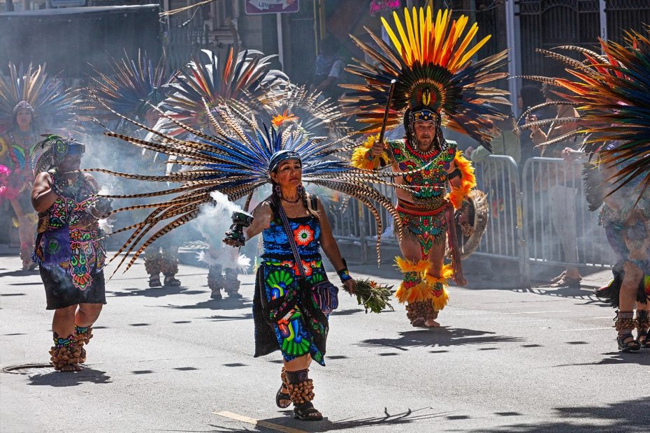 People in elaborate feathered headdresses and colorful traditional attire perform a cultural dance on a city street, with smoke in the background.
