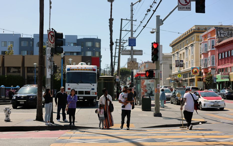 A group of people wait at a crosswalk on a city street with traffic lights, vehicles, and various buildings in the background on a sunny day.