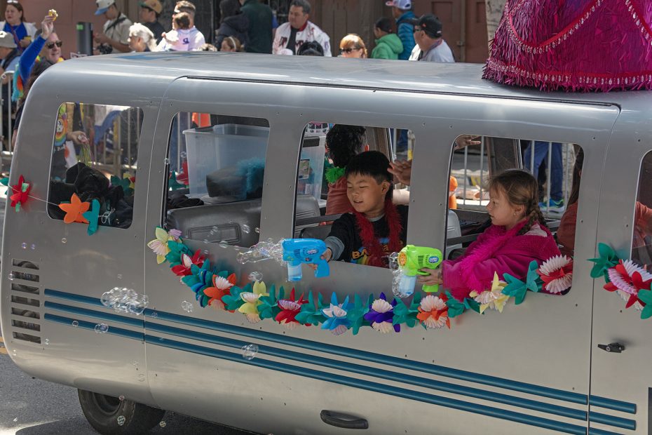 Two children inside a parade float decorated with colorful leis spray water guns out the window; bubbles float nearby as spectators watch from the street.