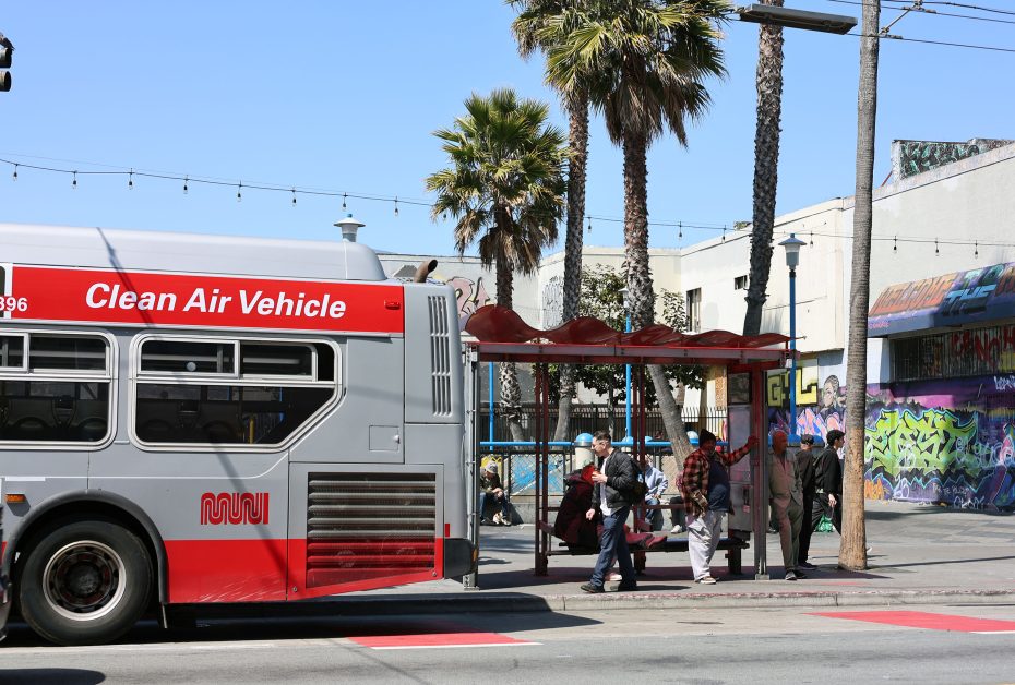 A city bus labeled "Clean Air Vehicle" stops at a bus shelter with people waiting and palm trees in the background on a sunny day.