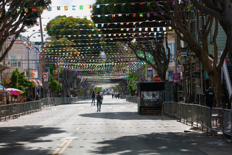 A mostly empty street lined with trees and barricades, decorated with hanging colorful flags, with a few people walking in the distance.
