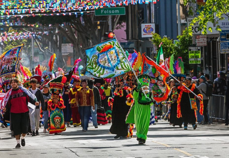 A group of people in colorful costumes and masks parade down a city street, carrying vibrant flags and banners; onlookers stand along the sidewalk.