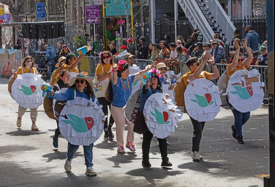 A group of people wearing fish-themed costumes parade down a street, surrounded by onlookers behind barricades.