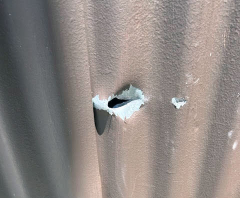 Close-up of a corrugated metal sheet with a jagged hole in the center and visible paint chipping around the damaged area, possibly caused by gunshots near Marshall Elementary.