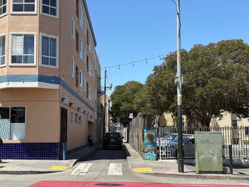 A narrow alley with a parked car between a peach building and a fenced lot with trees; street art is visible on a wall by the fence under clear blue sky.