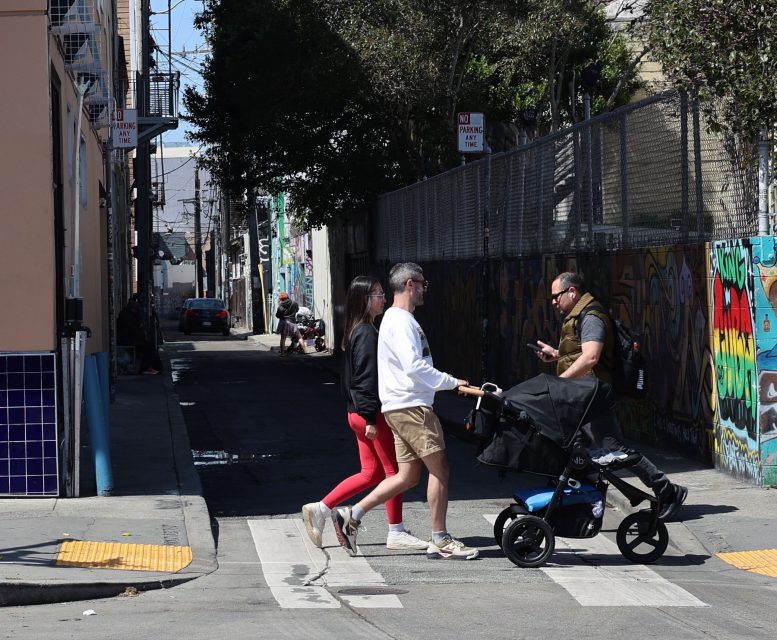 Three adults, one pushing a stroller, cross a street near a graffiti-covered alleyway in daylight.