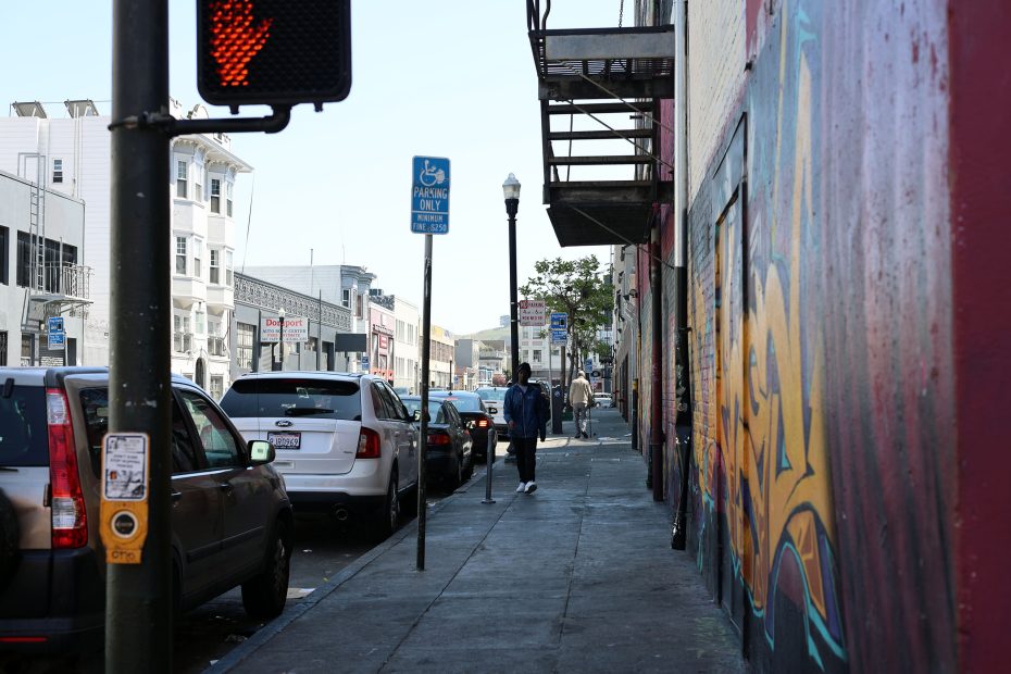 A city sidewalk with parked cars, a person walking, a traffic signal, and street art on a building wall.