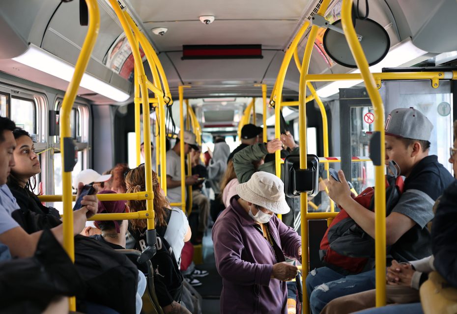 Passengers sit and stand inside a city bus, some wearing hats and masks, while others look at their phones or out the window. Yellow handrails and seats are visible throughout the bus.