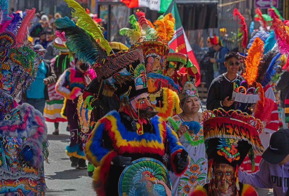 People in colorful, traditional costumes and feathered headdresses participate in a festive parade, with Mexican flags and onlookers in the background.