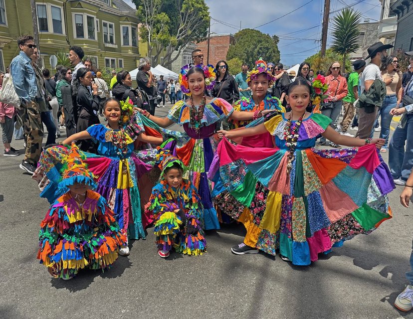 Five children in colorful traditional dresses pose in the street during a festival, surrounded by a crowd and houses in the background.