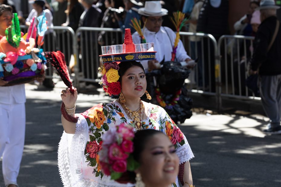 A woman in traditional Mexican attire and floral headdress participates in a parade, balancing a decorated tray on her head; others in festive dress are visible in the background.