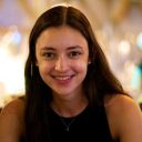 A young woman with long brown hair and a black top smiles at the camera in a softly lit indoor setting.