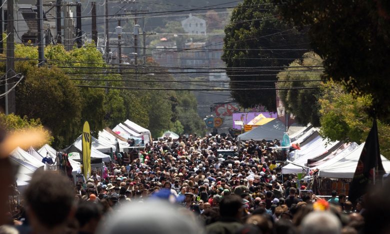 A large crowd walks down a city street lined with white vendor tents during an outdoor street fair or market.