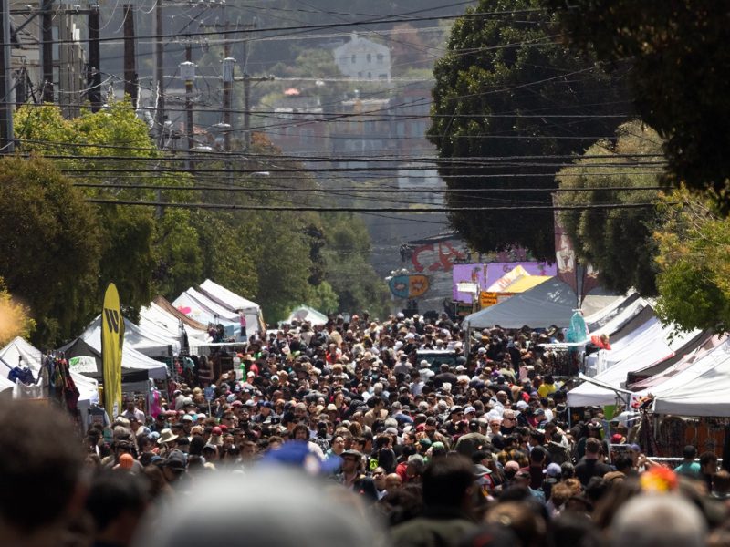 A large crowd walks down a city street lined with white vendor tents during an outdoor street fair or market.