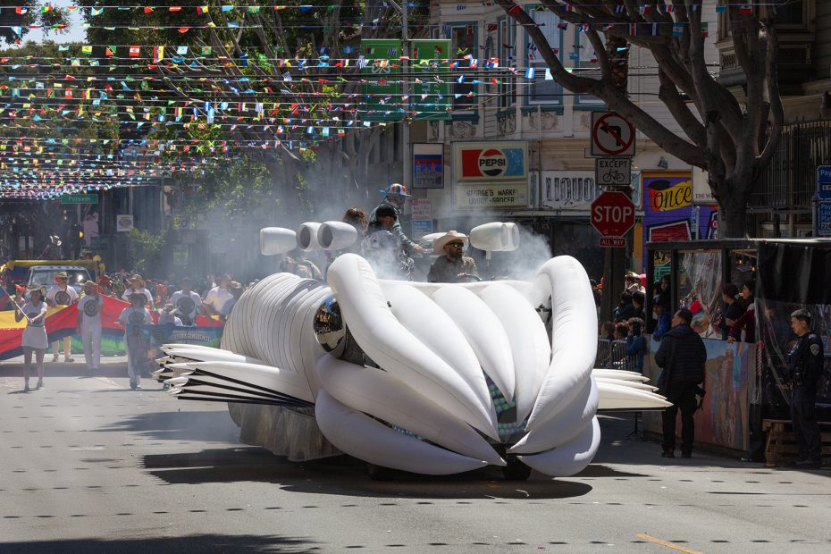 A large white inflatable float resembling a fish moves down a city street during a parade, with people inside and onlookers watching from the sidewalks.