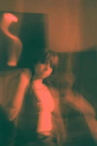 A blurred, motion-filled photo of a person standing indoors with indistinct features and a curvy abstract sculpture visible in the background. The image has a warm, muted color tone.