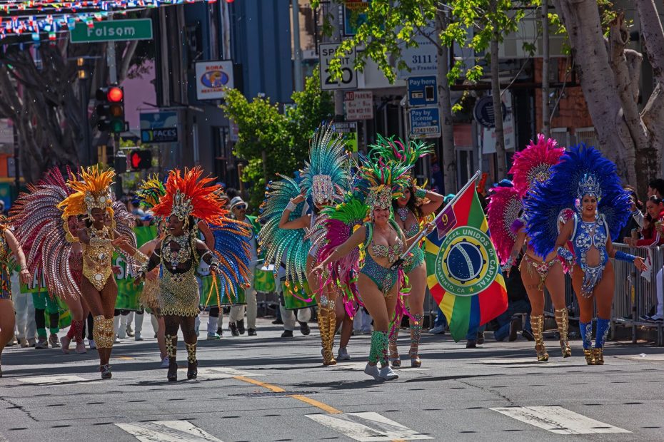 A group of dancers in colorful feathered costumes perform in a street parade, with one person holding a multi-colored flag in the background.