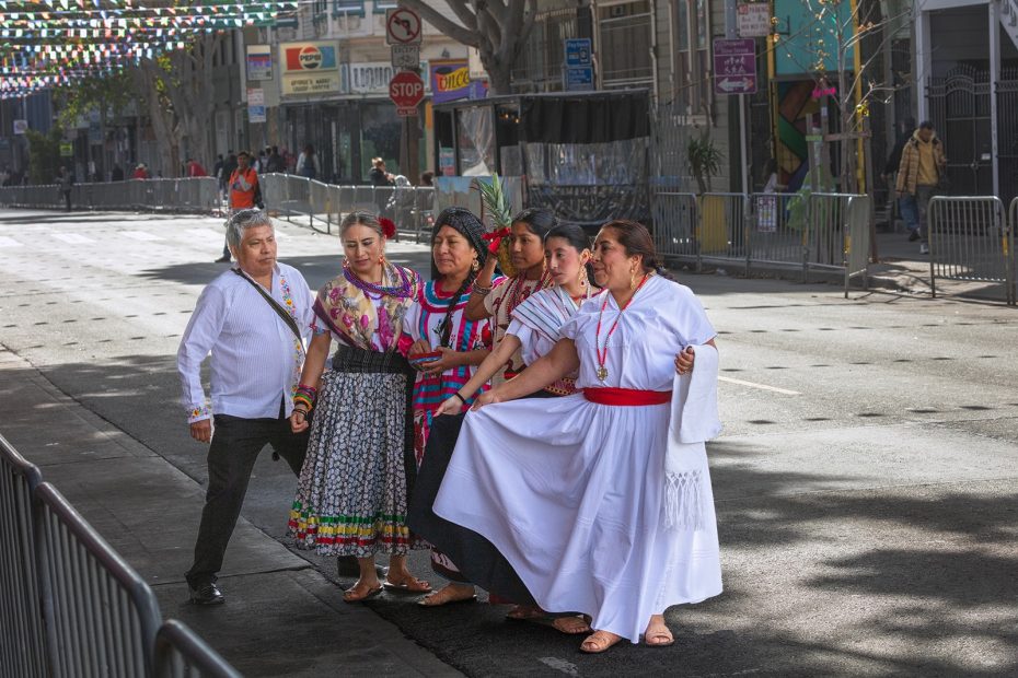 Five people dressed in traditional clothing pose together on a city street decorated with colorful banners, with metal barricades and buildings in the background.