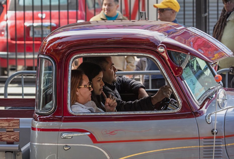 Three people are sitting inside a vintage red and silver pickup truck during a daytime event, with spectators and other vehicles visible in the background.