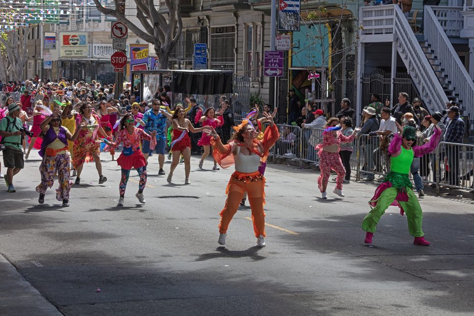 A group of people in colorful costumes dance in a parade on a city street, while spectators watch from behind metal barricades.