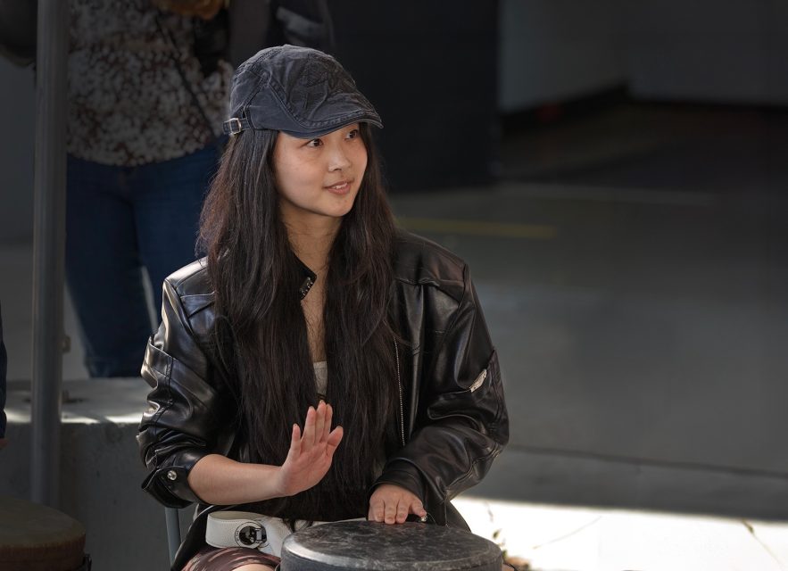 A person with long hair wearing a black jacket and cap is seated, playing a drum with one hand in an indoor setting.