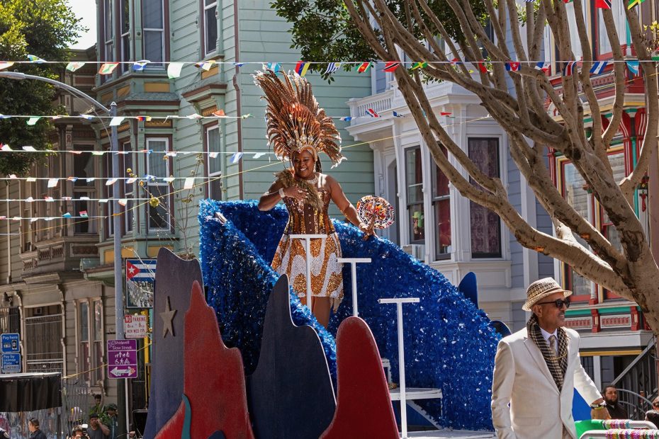 A person in a gold costume and headdress stands on a colorful parade float, while another person in a white suit walks in front. Urban street and row houses are in the background.