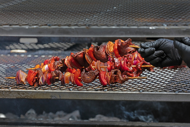 A gloved hand holds a skewer of grilled meat, onions, and peppers over a metal grill with several other skewers cooking.