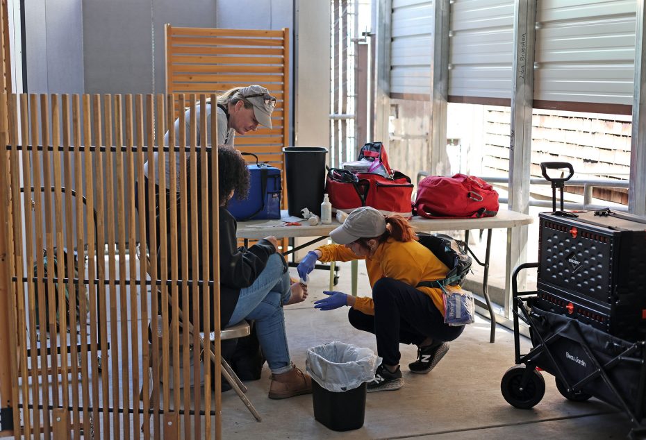 A seated person receives medical attention from a kneeling healthcare worker in a clinic setting, while another person observes. Medical supplies are visible on a table nearby.