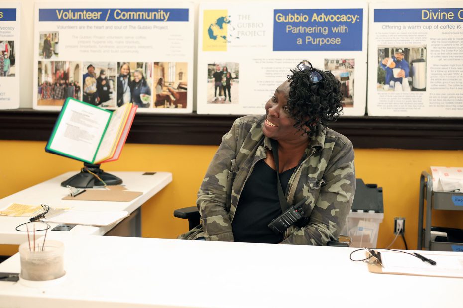 A woman in a camouflage jacket sits at a desk, smiling, with informational posters about community volunteer work displayed behind her.