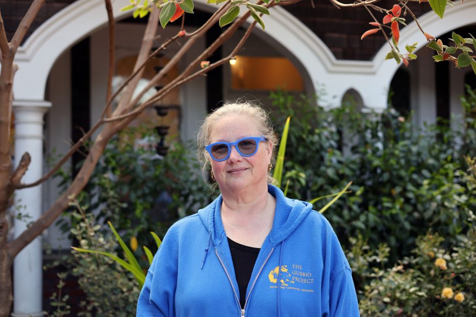 A woman wearing blue glasses and a blue hoodie stands outside in front of a house with plants and arched windows in the background.
