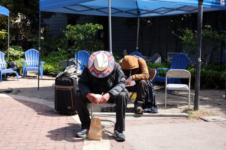 Two people sit hunched over on outdoor chairs under blue canopies, surrounded by bags and empty chairs. One wears a colorful hat, the other a hooded jacket.