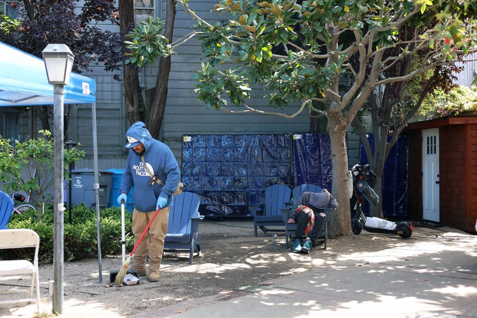 A person in a blue hoodie sweeps the ground near blue chairs and trees, while another person sits with their head down beside a shed and an electric scooter.