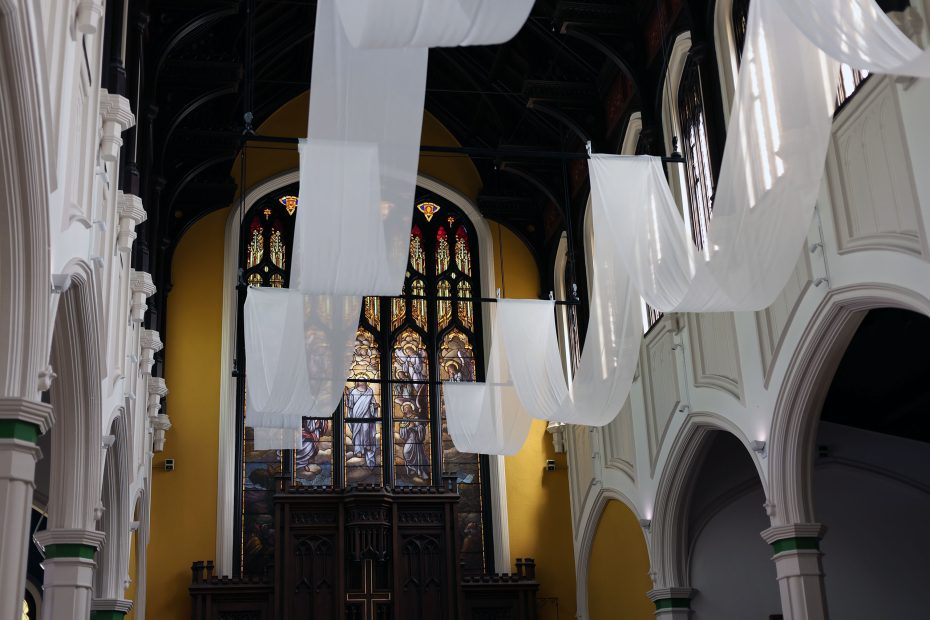 Interior of a church with arched white columns, stained glass windows, and white fabric draped from the ceiling.