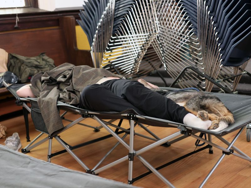 A person and a cat lie together on a cot in a room with wooden floors and stacked folding chairs in the background.