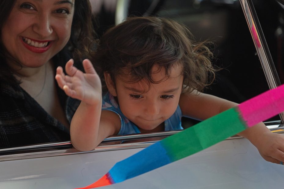 A woman smiles as a young child leans out of a car window, reaching toward a colorful ribbon outside the vehicle.