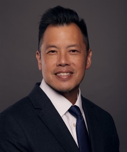 A man in a dark suit, white shirt, and navy tie poses against a plain dark background, smiling at the camera.