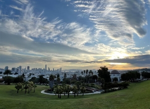 A city skyline is visible in the distance under a partly cloudy sky, with a green park and landscaped garden in the foreground at sunset.